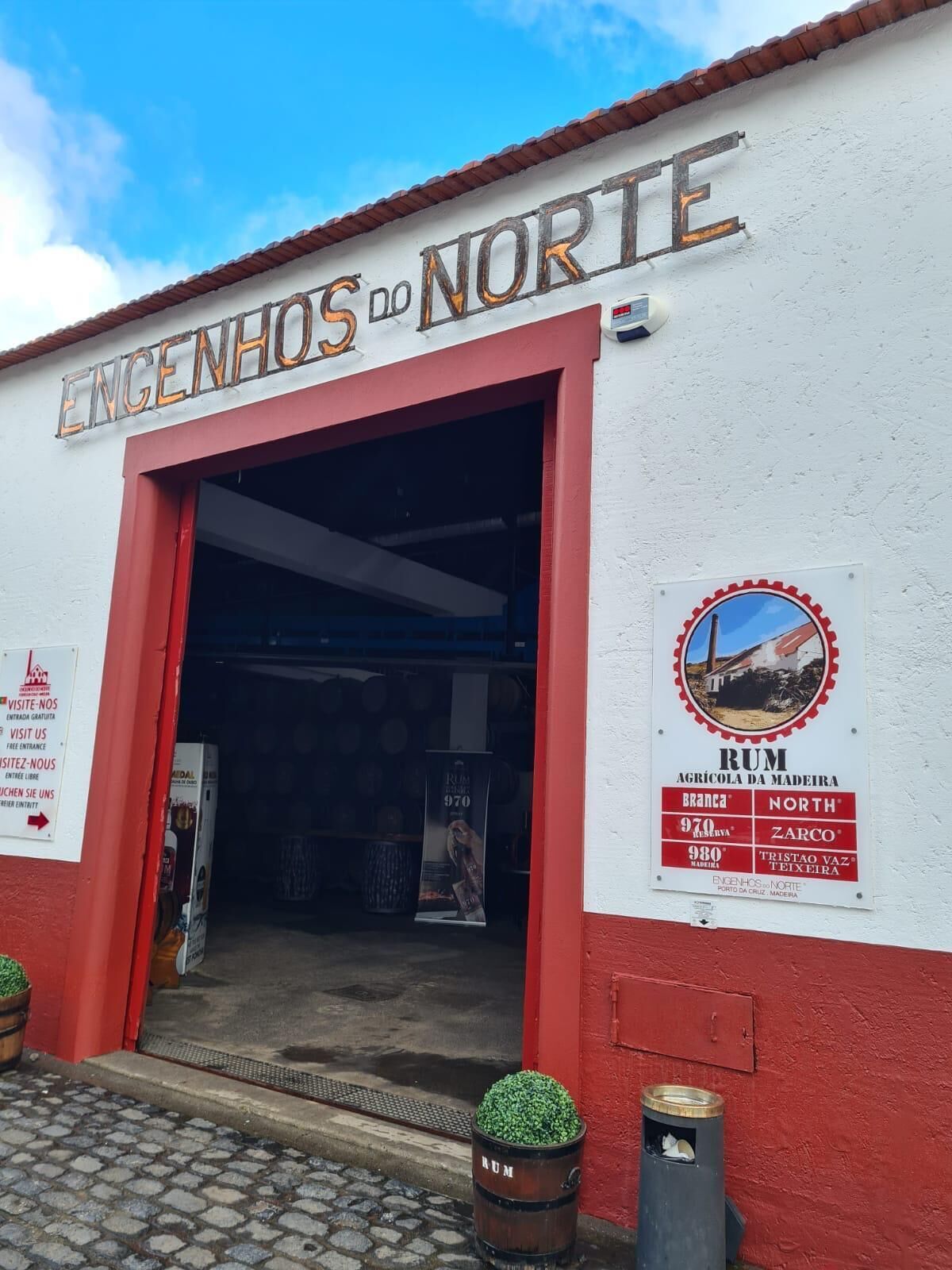 De ingang van Engenhos do Norte, een gebouw met een rood-witte gevel, met een grote open deur en een bord waarop rumproducten worden aangeboden.