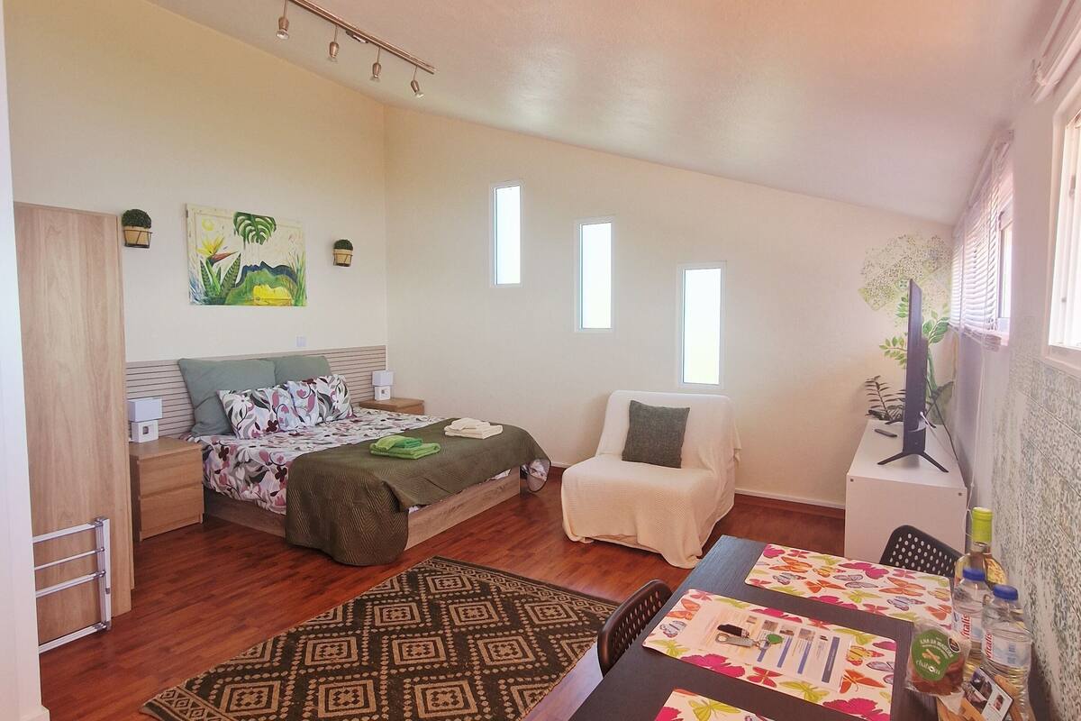 A spacious room with a bed, a chair, a table with a floral tablecloth, and a small plant on a side table.