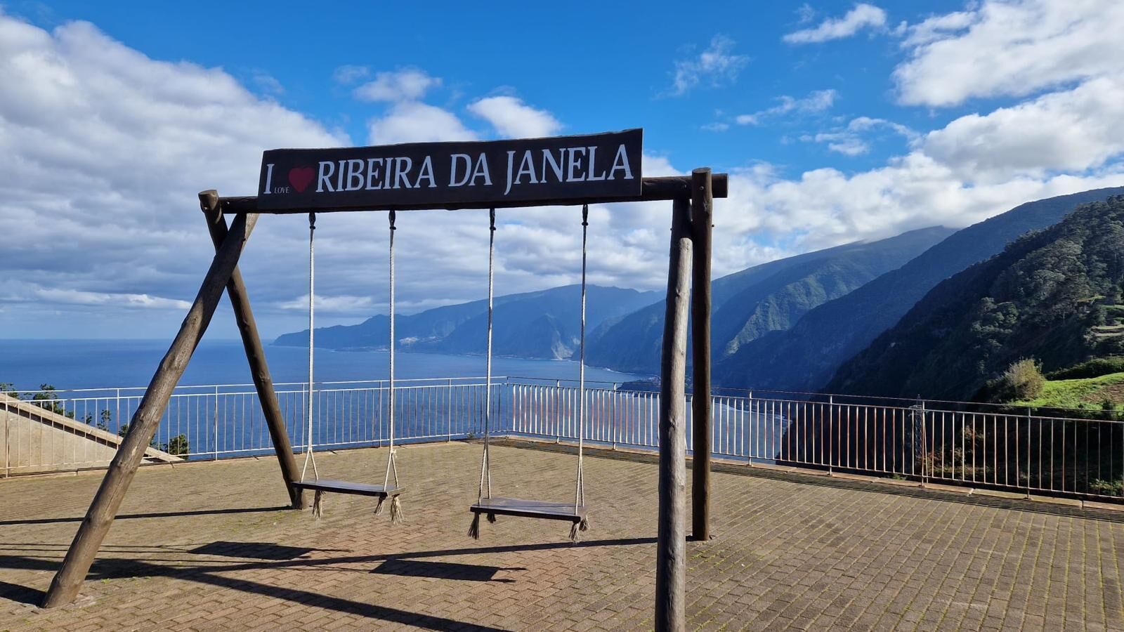 A swing set with a sign reading "RIBEIRA DA JANELA" in a scenic outdoor area with a view of mountains and the sea.