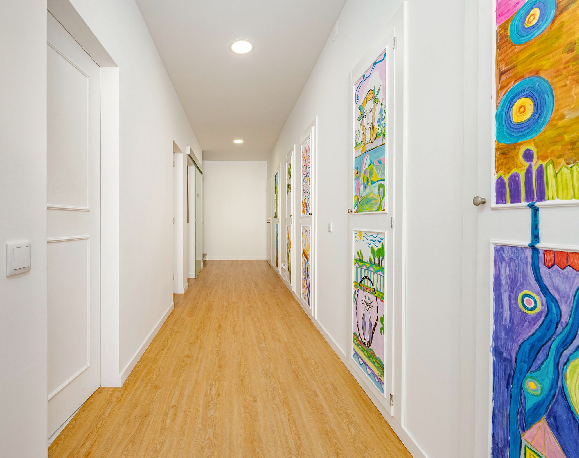 A hallway with wooden flooring and white walls, featuring colorful artwork on the doors.