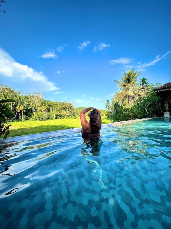 A clear pool with a person in the water, surrounded by lush greenery and a clear sky.