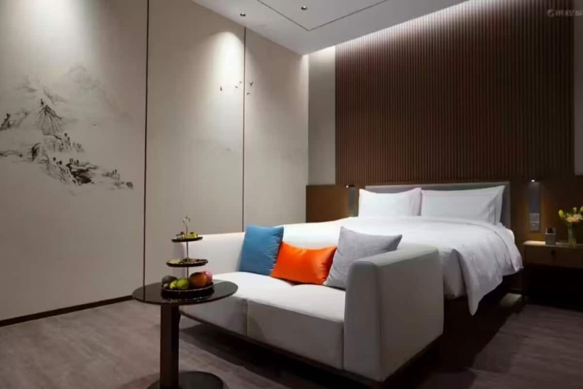A modern hotel room with a bed, a sofa, a small table with a fruit arrangement, and a wall with a vertical wooden panel.