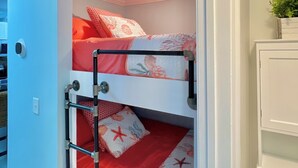 Una cama litera con colcha estampada en rojo y blanco, una escalera metálica y una pequeña planta en maceta sobre un estante.