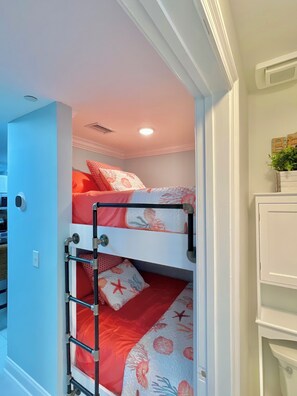 Una cama litera con colcha estampada en rojo y blanco, una escalera metálica y una pequeña planta en maceta sobre un estante.