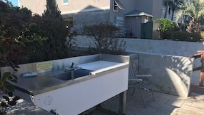 Un área de cocina al aire libre con pileta y encimera, rodeada por un muro de concreto y algunas plantas.