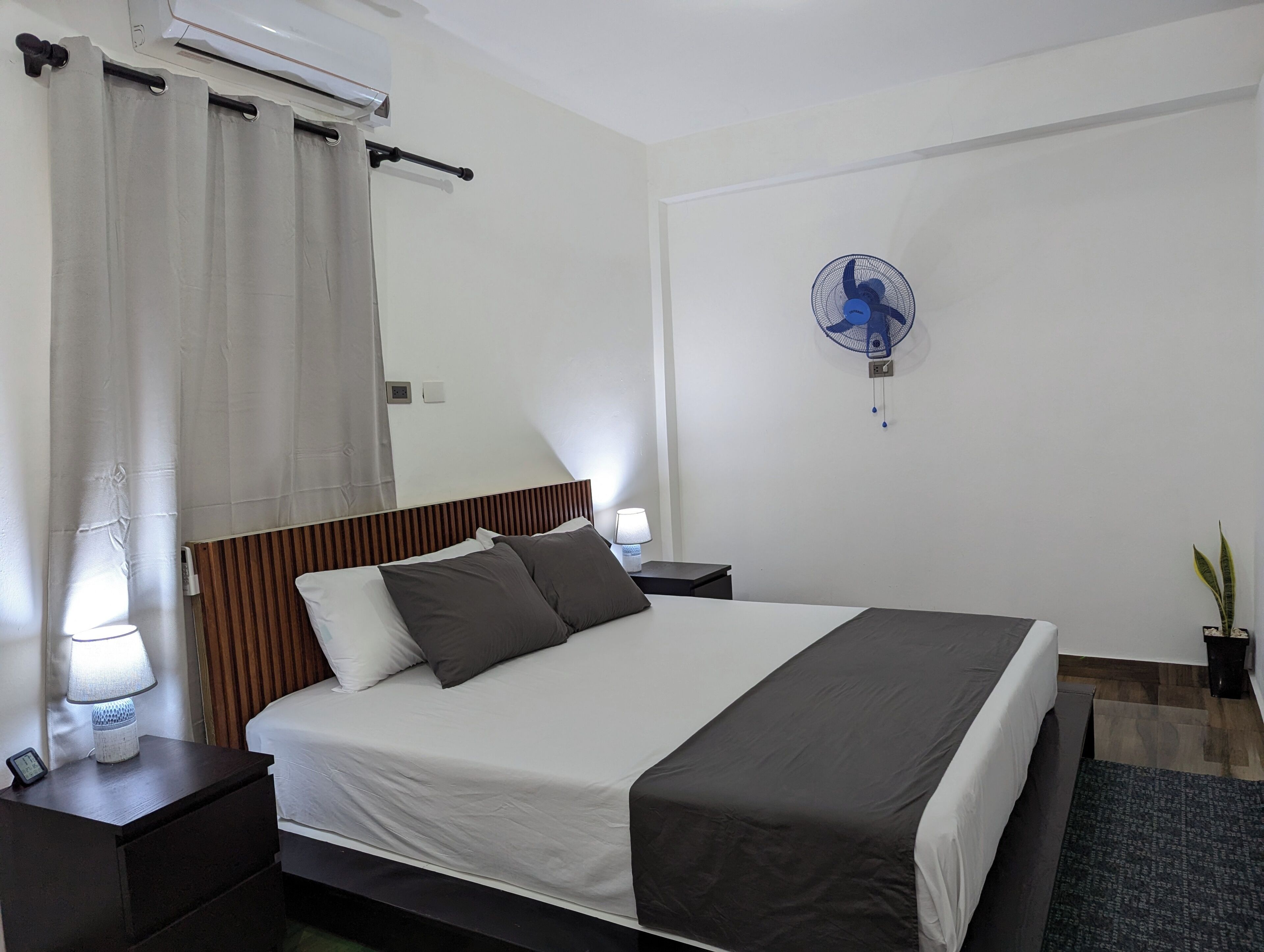 Una habitación de hotel con una cama grande, una mesita de noche con lámpara, aire acondicionado en la pared, ventilador de techo y una planta en maceta.