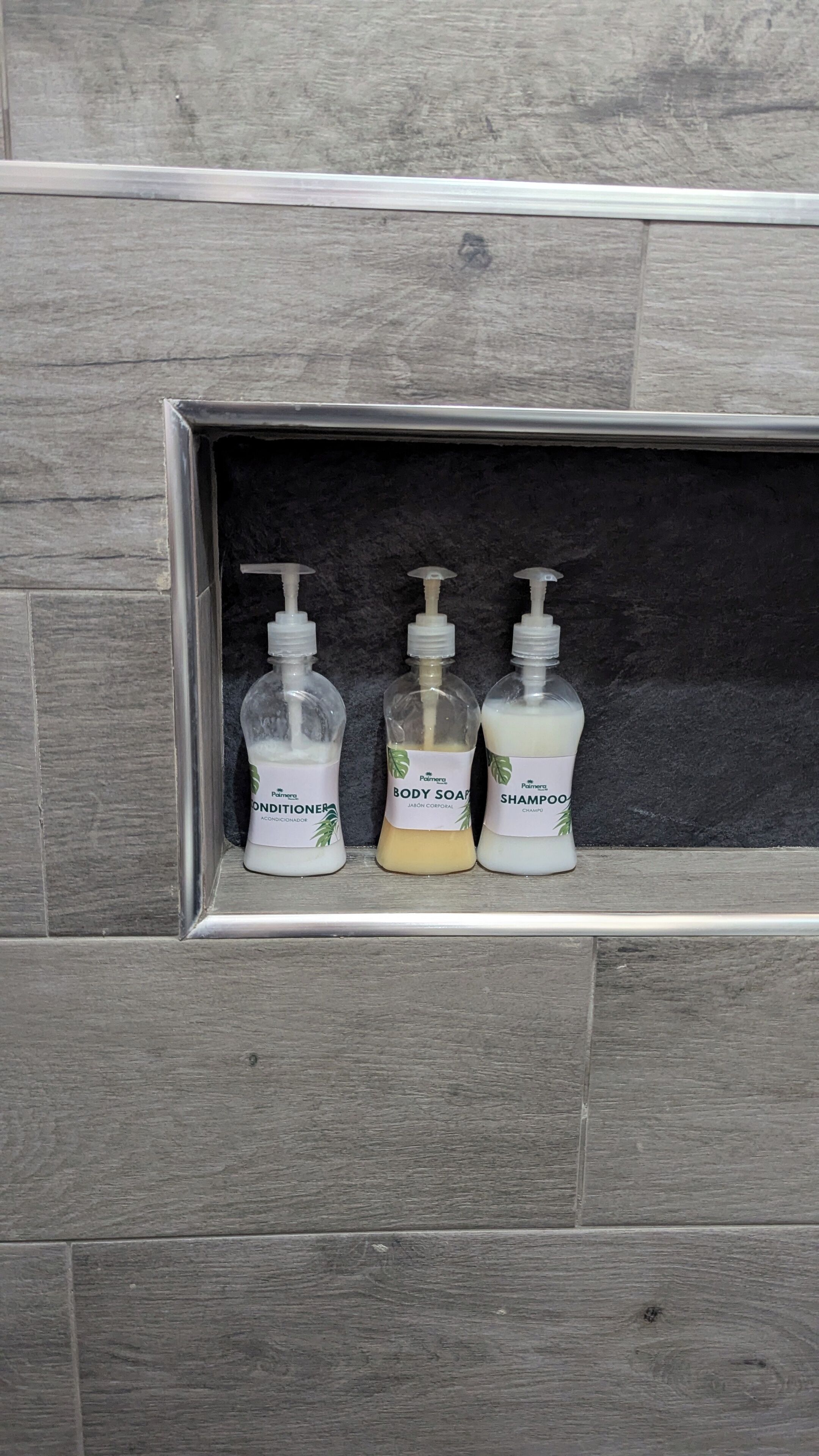 Three bottles of Palmera personal care products in a shelf against a grey tiled wall.