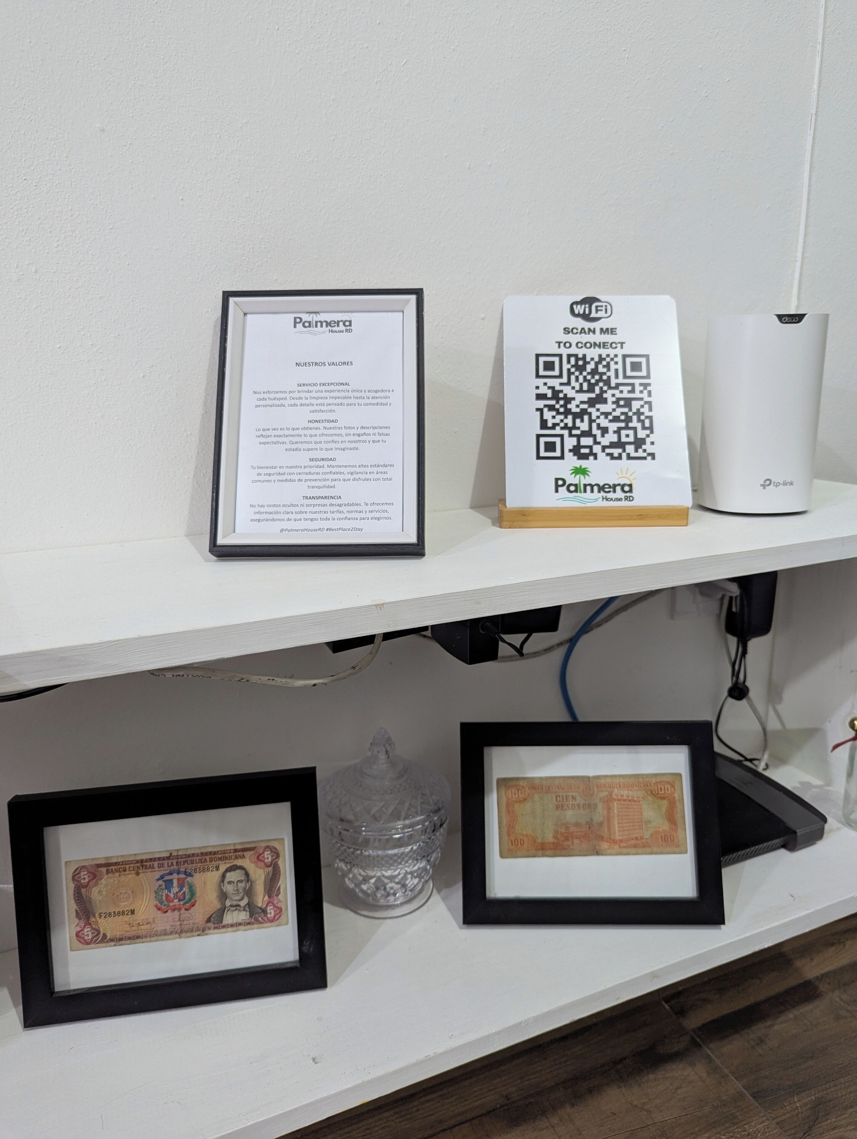 Un documento enmarco, un letrero con un código QR, un router Wi-Fi y dos billetes enmarcados sobre una estantería.