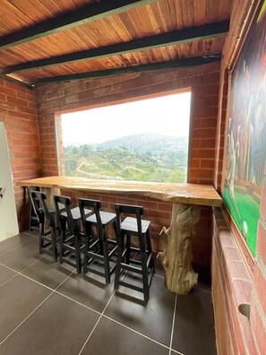 A rustic dining area with a wooden table and chairs, a large window overlooking a hilly landscape, and a mural on the wall.