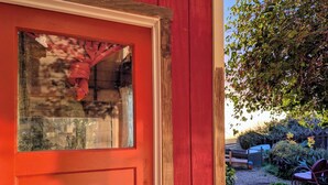 A red door with a glass panel, a metal door lock, and a view of a garden with plants and a wooden fence. - Hills Cottage with Panoramic Views & Hot Tub. Pet Friendly! (Oakland)