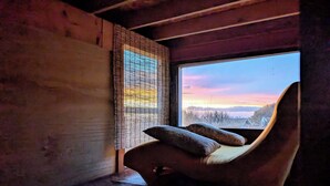 A cozy room with a wooden interior, a large window with a view of a sunset, and a unique curved chair with cushions. - Hills Cottage with Panoramic Views & Hot Tub. Pet Friendly! (Oakland)