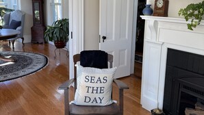 A cozy living room with a fireplace, a clock, and a chair with a cushion that says "SEAS THE DAY". - Harbor Water View. Downtown. Beaches & waterfront playground +Cape Cod gateway! (Plymouth)