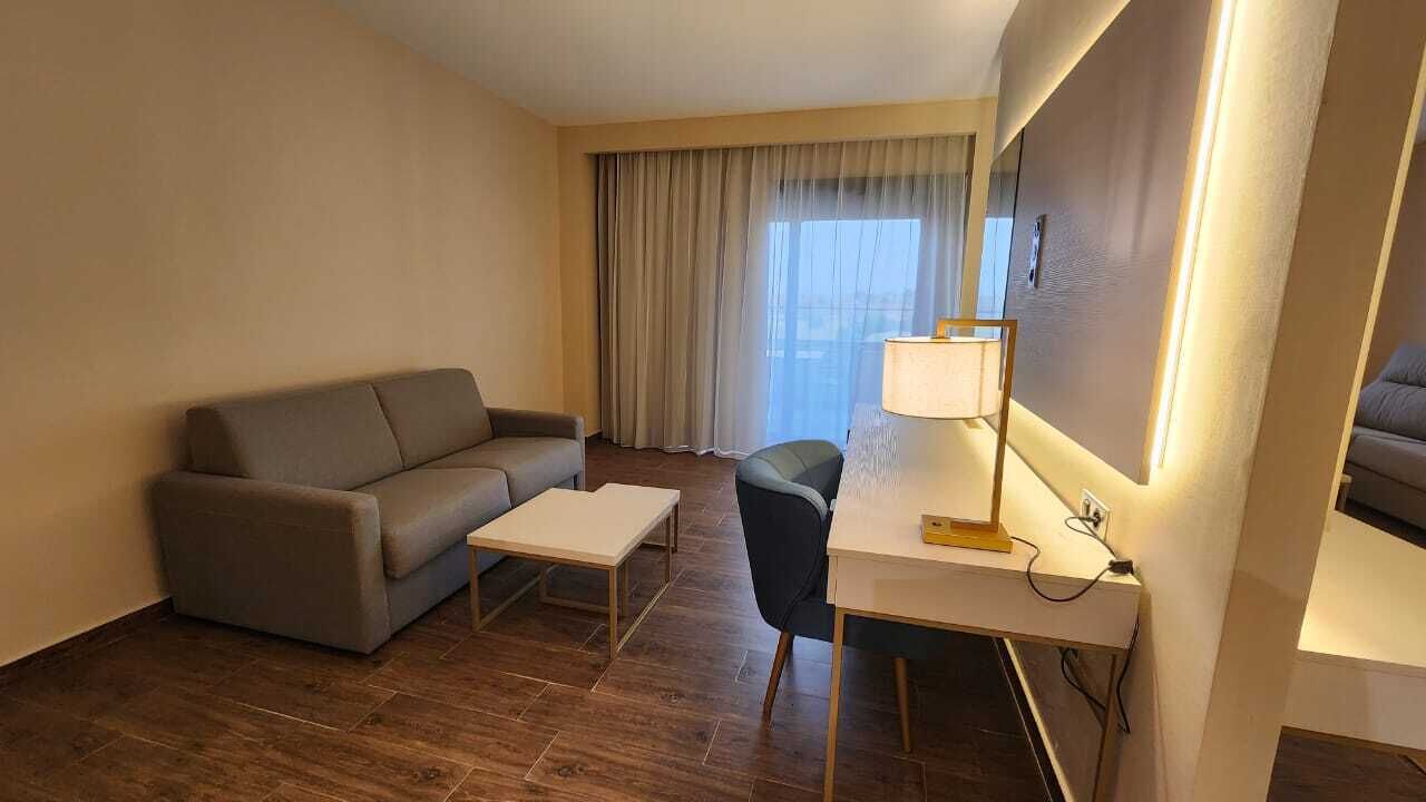 Une chambre d’hôtel avec un canapé, une petite table, un bureau avec une lampe et une grande fenêtre avec des rideaux.