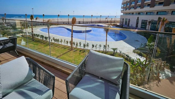 Balcón con mobiliario de mimbre con vistas a una piscina y una playa.