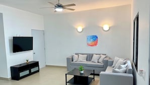 Una sala moderna con un sofá gris, una mesa de centro, un televisor de pantalla plana, un ventilador de techo y dos luces fijadas en la pared.