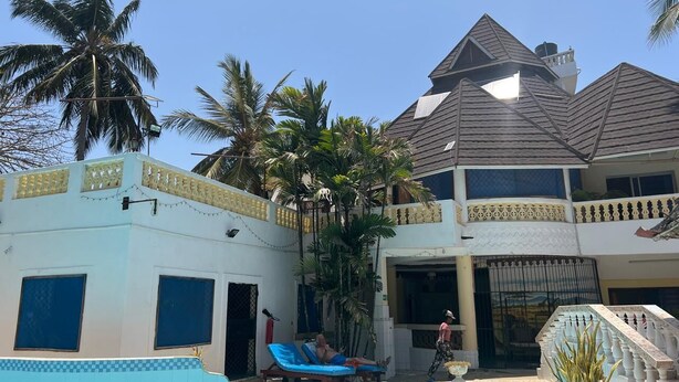 Un bâtiment à deux étages avec une piscine, des palmiers et une personne qui se promène.