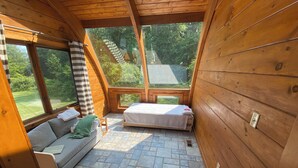 Acogedora habitación tipo cabaña con interior de madera, suelo de baldosas de piedra, cama, sofá y ventanas amplias con vistas al exterior.