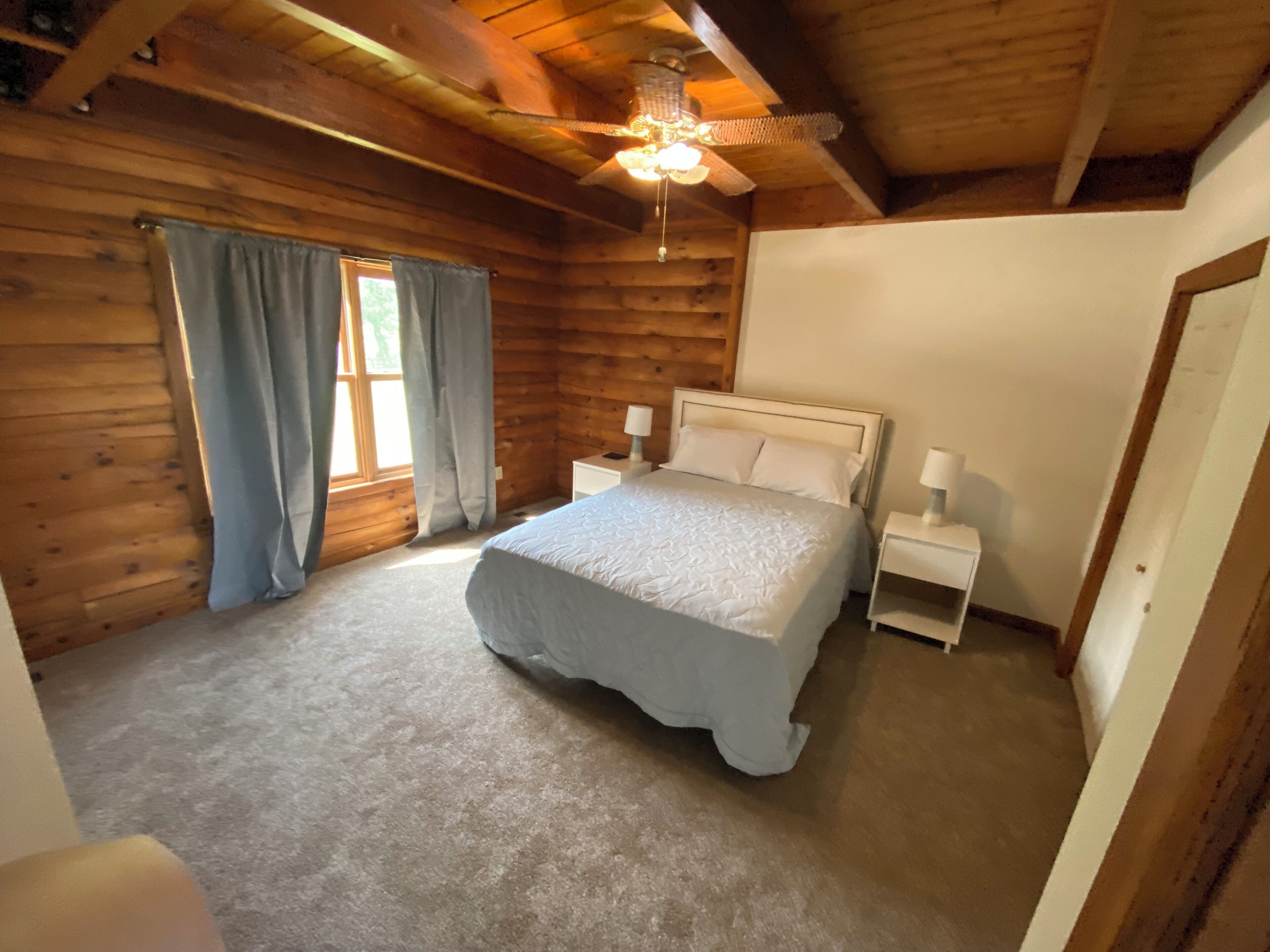 Un dormitorio de cabaña de madera con cama, dos mesitas de noche, ventilador de techo y ventana con cortinas.