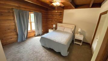 Un dormitorio de cabaña de madera con cama, dos mesitas de noche, ventilador de techo y ventana con cortinas.