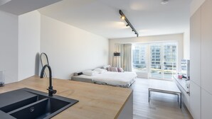 A modern hotel room with a kitchenette, a bed with white bedding, a large window with curtains, and a view of buildings. - Modern Studio With sea View (Middelkerke)