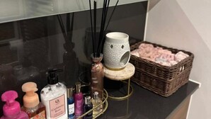 A bathroom vanity with a black countertop, a white mug with a geometric pattern, a wicker basket with pink items, and various toiletries. - 1 Bed Apartment Sleeps 2 - Free Parking - (Dartford)