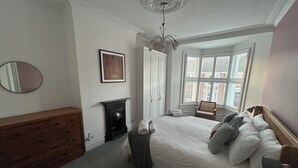 A bedroom with a large bed, a dresser, a mirror, a fireplace, and a window with blinds.