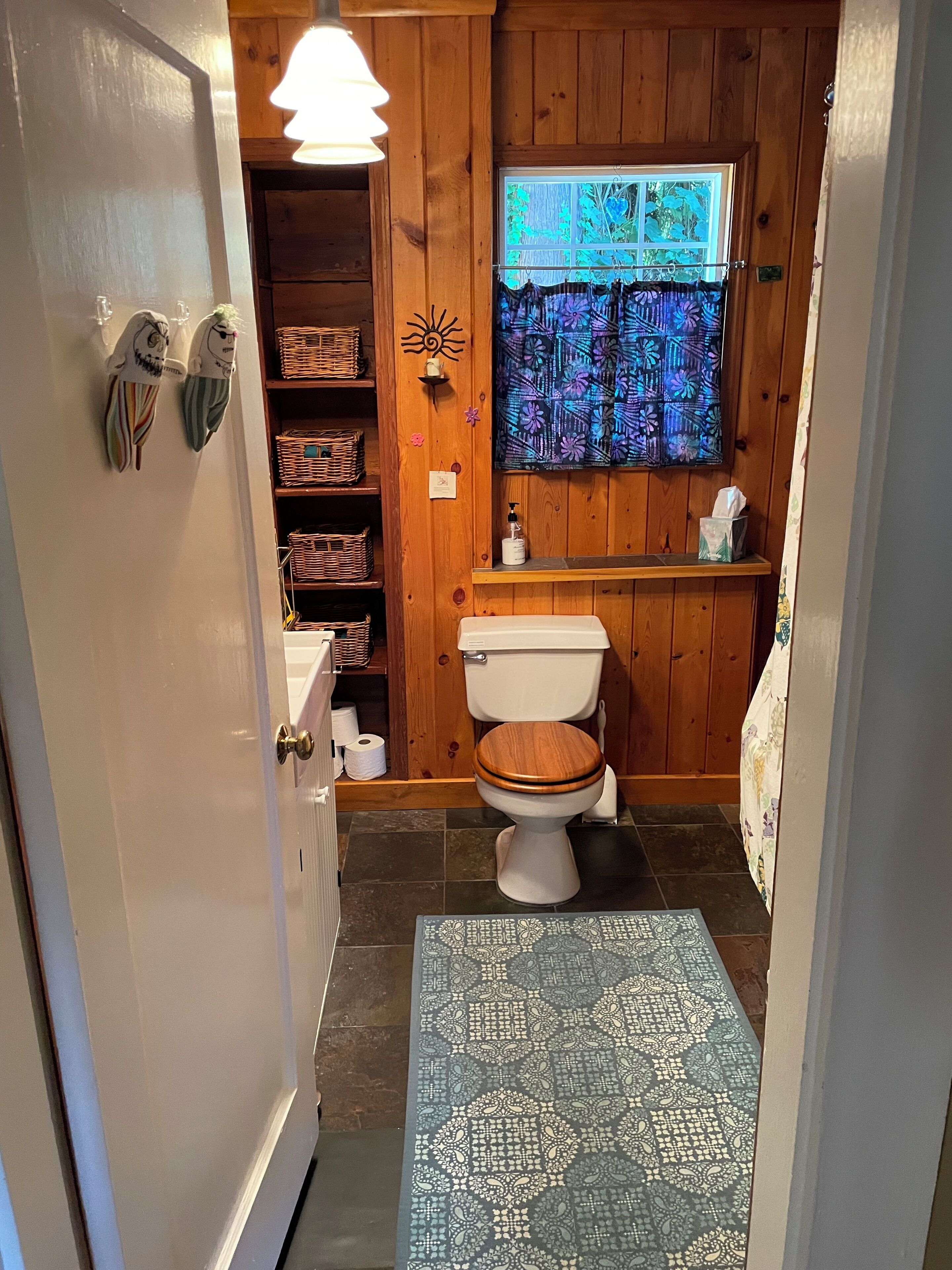 Een badkamer met een houten wastafelmeubel, een toilet, een raam met bloemenmotief en een vloerkleed met dessin.