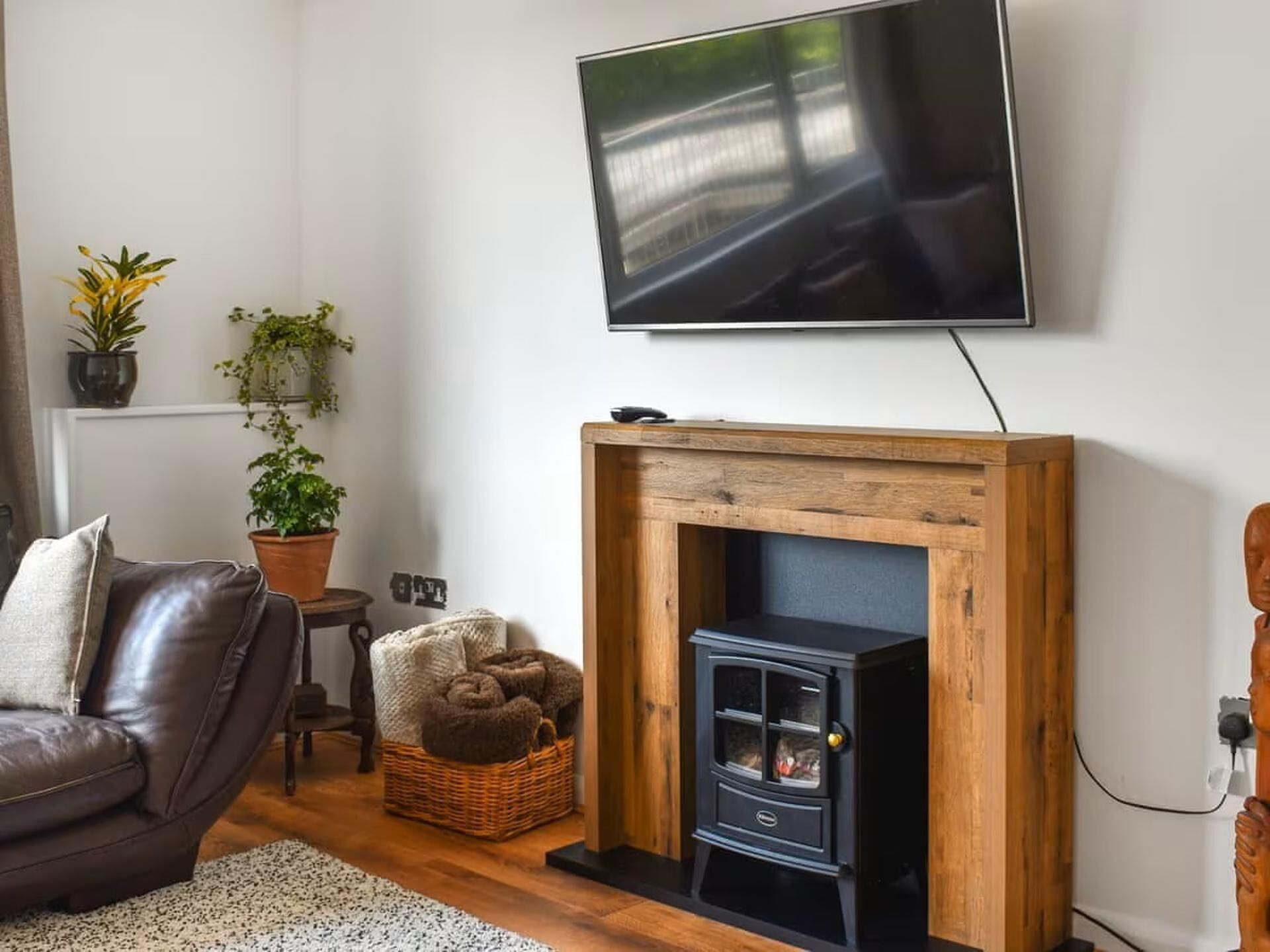 A living room with a black wood-burning stove, a leather sofa, a TV mounted on the wall, and potted plants.
