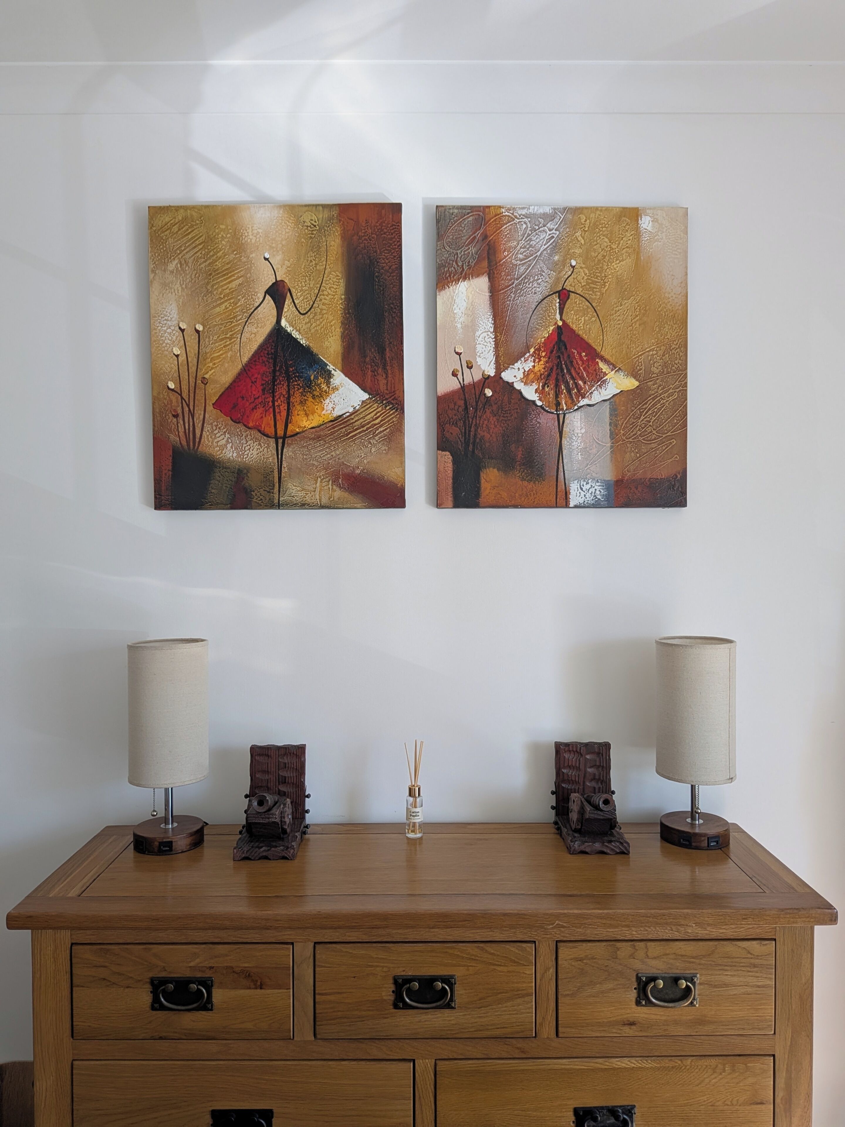 A wooden dresser with two wall-mounted paintings, a small vase with reed diffusers, and two table lamps.