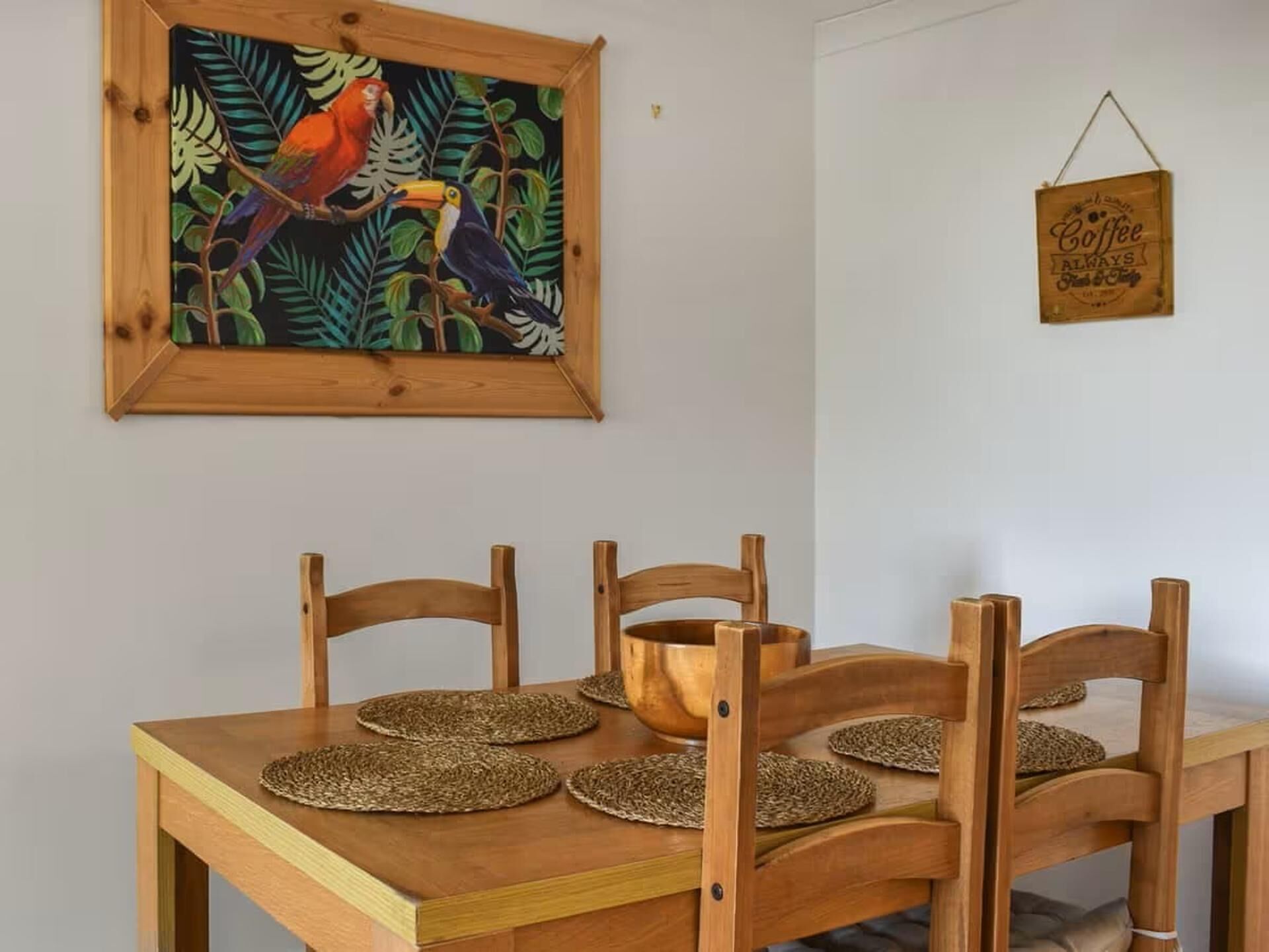 Une table de salle à manger en bois, agrémentée de sets de table tressés et de chaises, un tableau encadré représentant un oiseau tropical et une enseigne suspendue sur laquelle on peut lire : « Café toujours frais et chaud ».