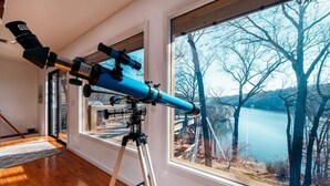 A telescope on a tripod in a room with large windows overlooking a lake and trees. - Winding Down at Lake Ann - Stunning Views Await! (Bella Vista)