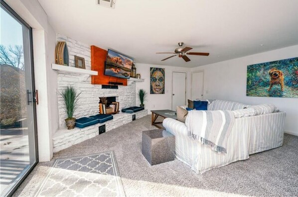 A modern living room with a stone fireplace, a ceiling fan, and a large painting of a dog. - Winding Down at Lake Ann - Stunning Views Await! (Bella Vista)