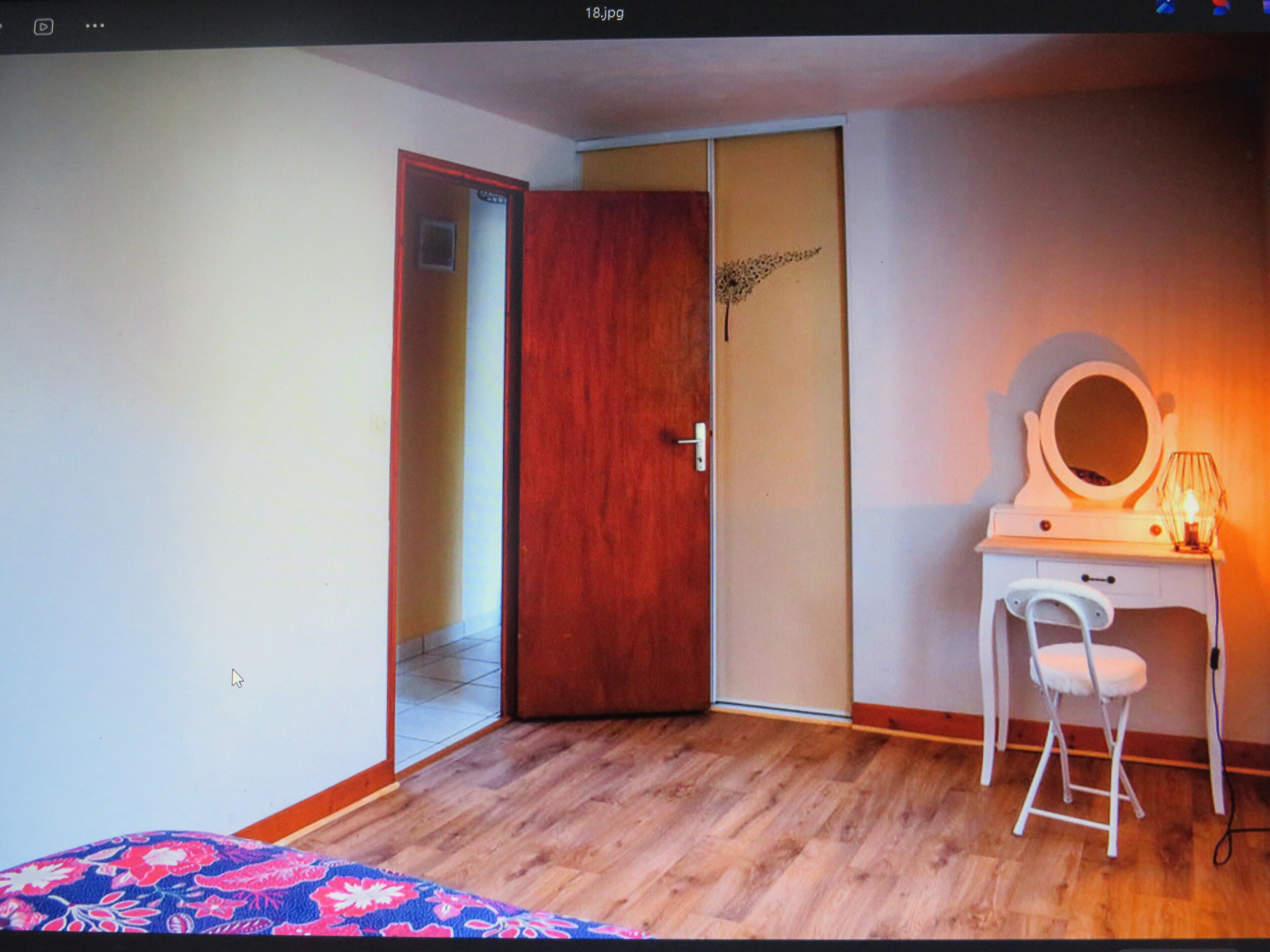 A room with a wooden floor, a white desk with a mirror and lamp, a white chair, and a door leading to another room.