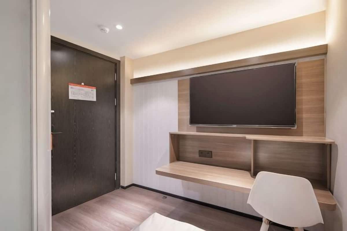 A modern hotel room with a dark wooden door, a mounted flat-screen TV, built-in shelves, and a white chair.
