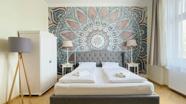 A double bed with a grey upholstered headboard, two bedside tables with lamps, and a patterned tapestry on the wall.