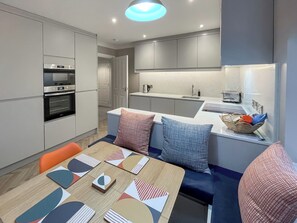 Eine moderne Küche mit eingebauten Geräten, ein Essbereich mit einem Holztisch und bunten Untersetzern sowie eine gemütliche Sitzecke mit gemusterten Kissen.