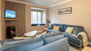 A modern living room with a sofa, a small table, a TV, a fireplace, and a window with a view of buildings. - 3 bedroom accommodation in Whitby (Whitby)