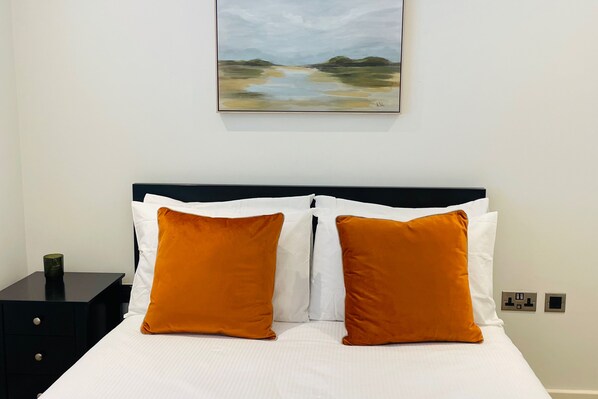 A neatly made bed with white and orange pillows, a black headboard, and a painting of a landscape above it. - Stylish Modern Apartment in St Albans City (St Albans)