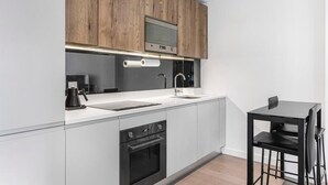 A modern kitchen with wooden cabinets, a built-in oven, a sink, and a small dining area with a table and chairs. - Stylish Modern Apartment in St Albans City (St Albans)