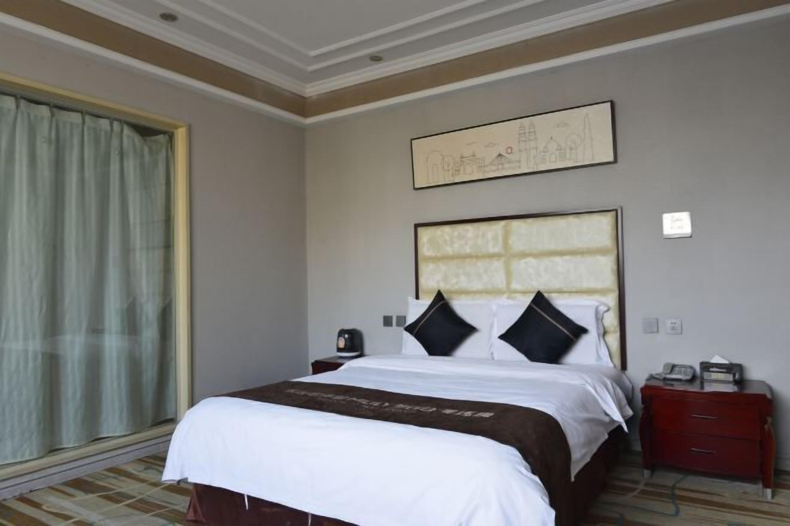 A hotel room with a bed, a nightstand, a window with curtains, and a wall-mounted artwork.
