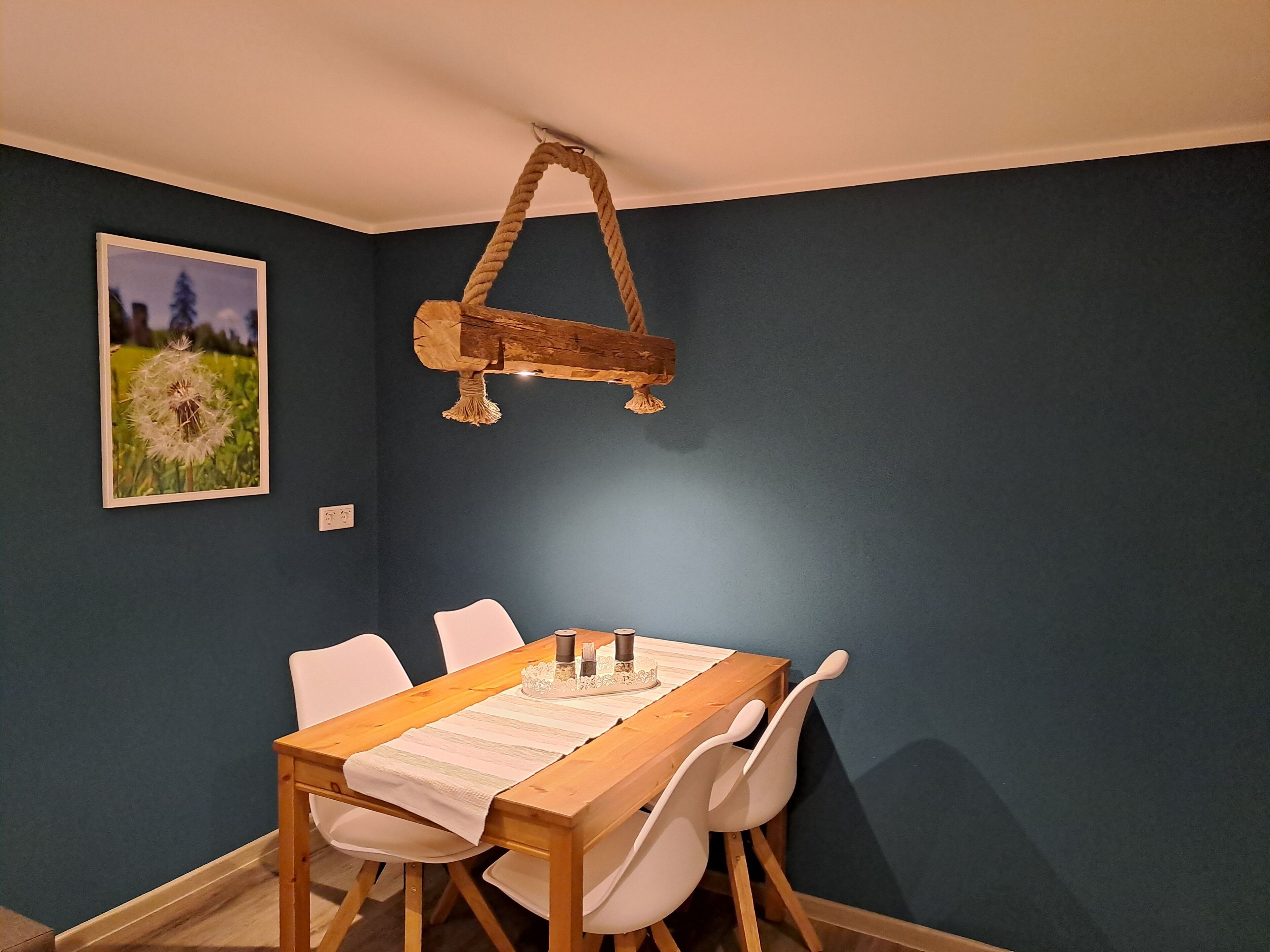 A dining area with a wooden table and white chairs, a hanging wooden light fixture, and a framed picture on the wall.