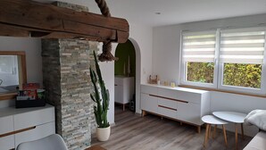 Una sala moderna con pared de piedra, vigas de madera, un ventanal con persianas, un sofá blanco, una mesa de comedor y una planta de cactus.