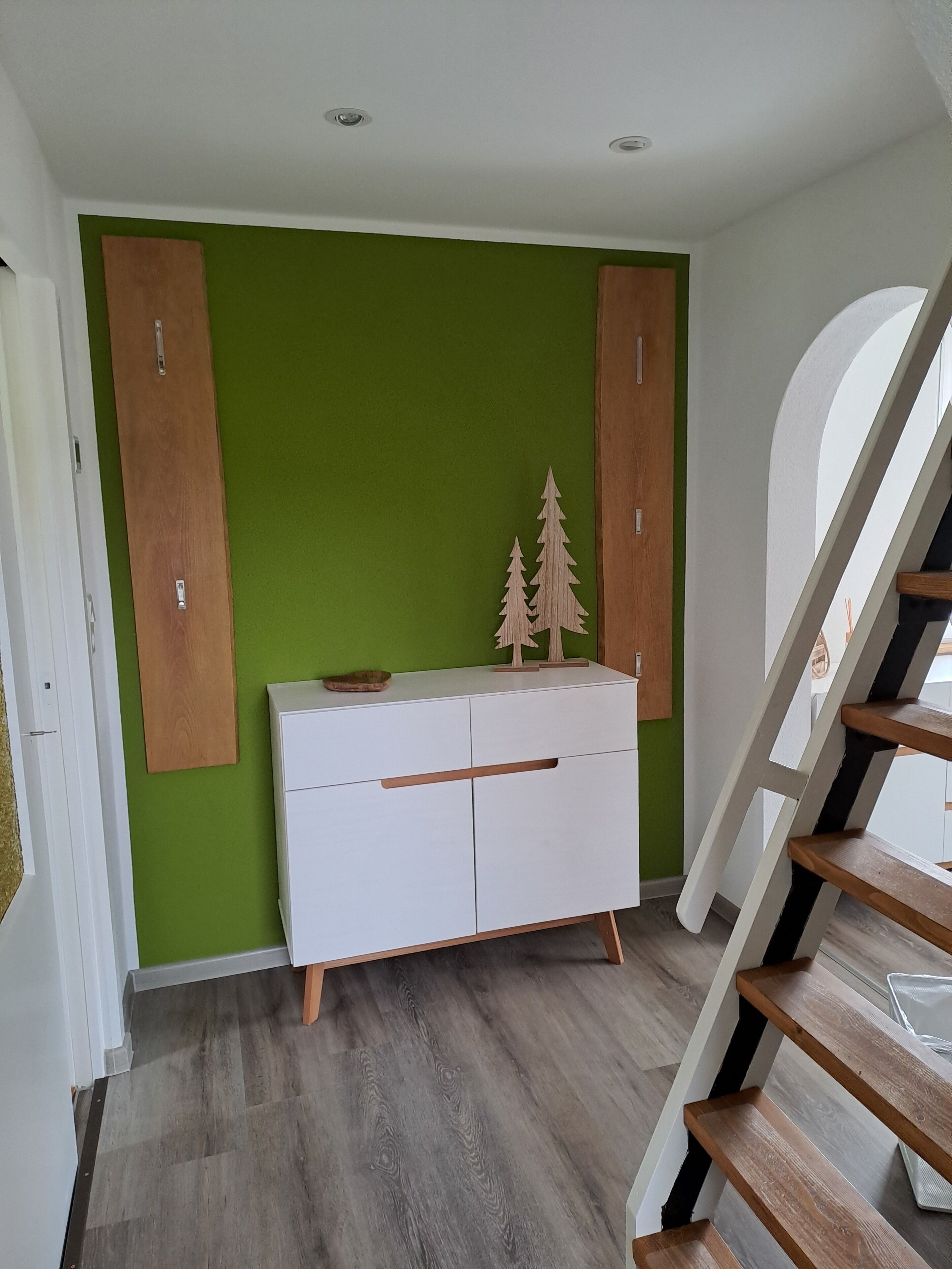A room with a white dresser against a green wall, wooden cabinets, a staircase, and a small wooden tree decoration on the dresser.