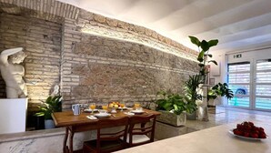 A dining area with a stone wall, a statue, and a table set for two with fruit and drinks. - Colosseum Loft | Monti ArtDesignApartments (Roma)