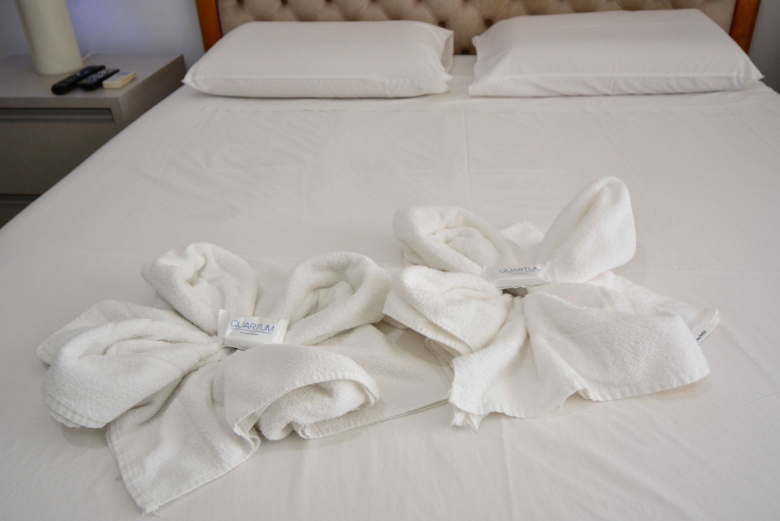 A bed with two towels folded into bows, a remote control, and a lamp on a bedside table.