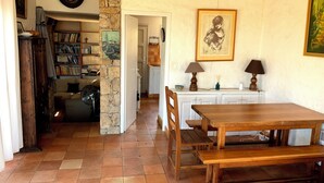 Uma sala de estar acolhedora com uma mesa de jantar de madeira, uma estante e uma cozinha visível através de uma porta aberta.