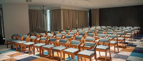 Un salón de conferencias con filas de sillas de madera y cojines azul claro, una alfombra estampada y una pantalla grande oscura en una pared.