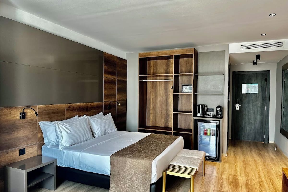 Ett modernt hotellrum med en stor säng, sängbord, en inbyggd garderob, en minibar och en dörr till badrummet.