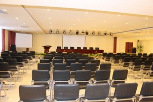 Un salón de conferencias con filas de sillas, un atril, una pantalla y un escenario.