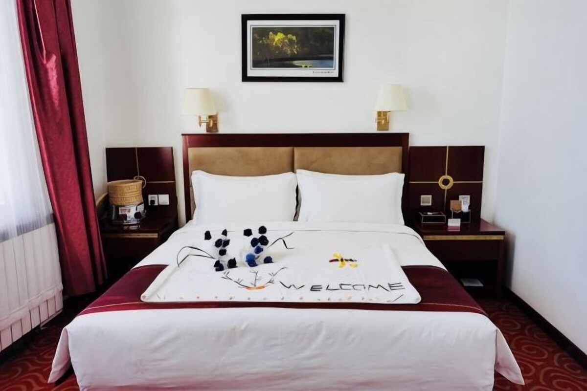 Ein Hotelzimmer mit einem Bett, zwei Nachttischen, einer Lampe, einem gerahmten Bild an der Wand und einer Fußmatte mit floralem Muster.